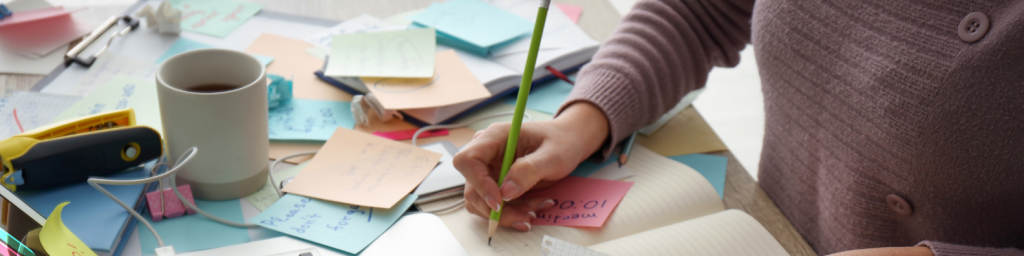 Marketing-Freelance am Arbeitsplatz: Person schreibt Notizen im Planer, umgeben von bunten Post-its, Laptop, Kaffee und Marketing-Ideen auf einem strukturierten Schreibtisch.