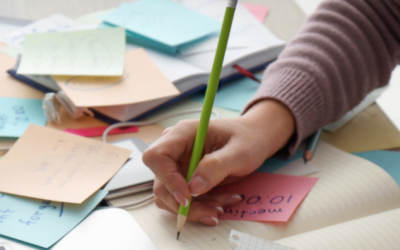 Marketing-Freelance am Arbeitsplatz: Person schreibt Notizen im Planer, umgeben von bunten Post-its, Laptop, Kaffee und Marketing-Ideen auf einem strukturierten Schreibtisch.